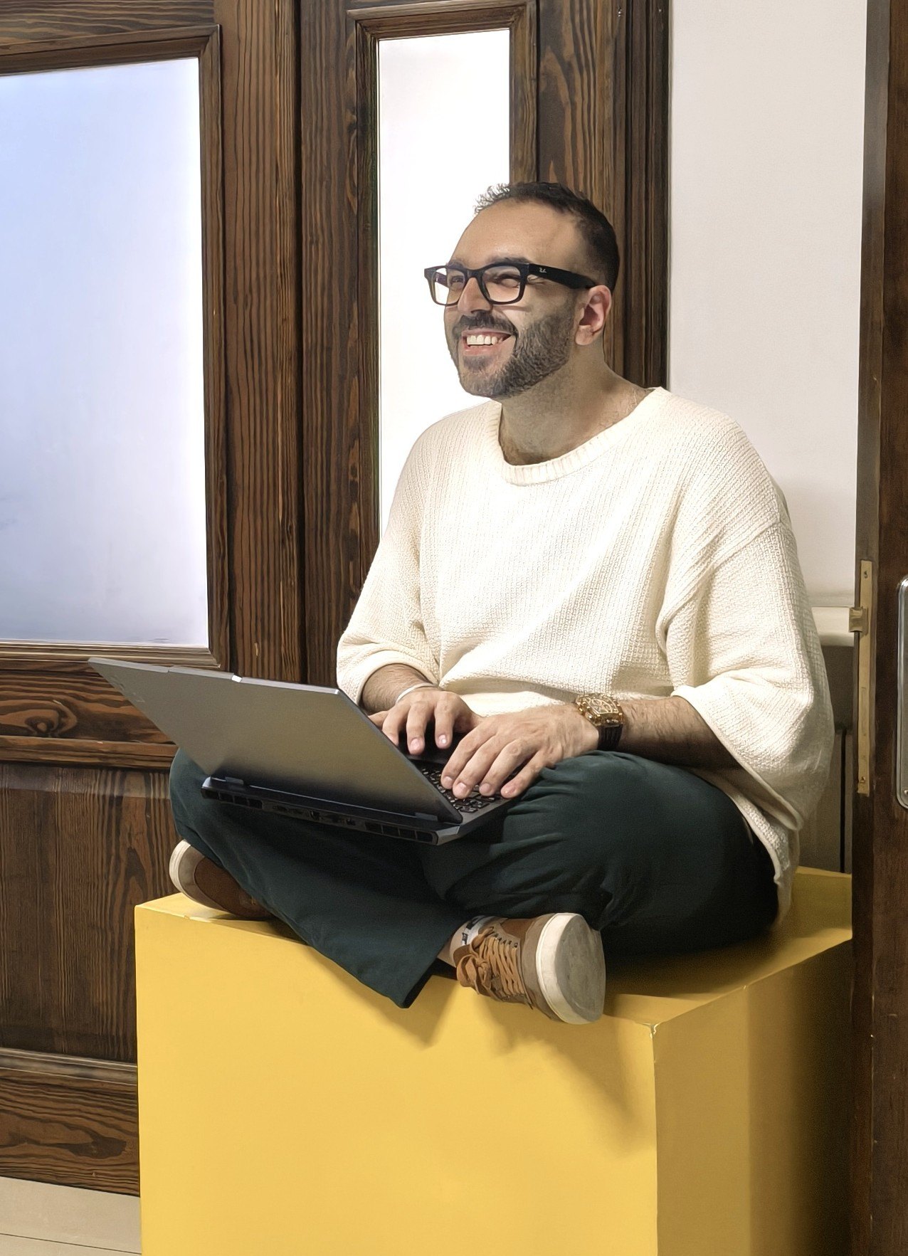 Kamel laughing, sitting cross-legged on a yellow cube with a laptop